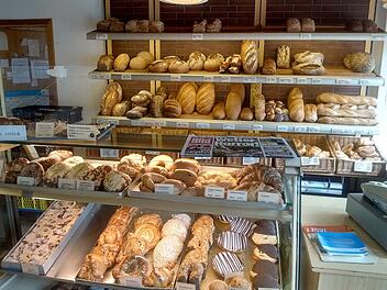 Götz-Brot: Fränkische Großbäckerei stellt nach 135 Jahren Betrieb ein Ein Bäckereigeschäft zeigt gefüllte Regale mit vielfältigem Gebäck, Broten und Brötchen. Im Vordergrund liegt eine Zeitung. Leider wird die Bäckereikette bald schließen müssen.