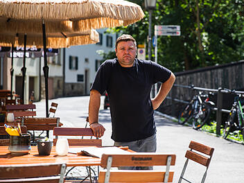 Bamberg: "Weinfass"-Gastronom ärgert sich über die Stadt Ein Mann steht draußen vor einem Restaurant mit leeren Tischen und Schirmen. Im Hintergrund ist eine gesperrte Brücke. Fahrräder lehnen am Geländer.