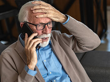 An elderly man with glasses and a beard, wearing a blue shirt and beige cardigan, looks distressed while talking on the phone, holding his head in concern An elderly man with glasses and a beard, wearing a blue shirt and beige cardigan, looks distressed while talking on the phone, holding his head in concern