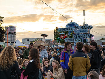 Bamberg: Kontakt-Festival vor ungewisser Zukunft - "offen, wie es weitergeht" Das Bild zeigt ein lebhaftes Festival mit vielen Besuchern, Essenständen und einer Außenbar. Ein Schild weist auf verschiedene Attraktionen hin. Der Himmel ist teils bewölkt beim Sonnenuntergang.